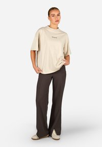 Bežs pārmērīgs T-krekls ar īsām piedurknēm un diskrētu logotipu, apvienots ar brīviem tumši pelēkiem biksēm ar sānu griezumiem un krēmkrāsas kedām.