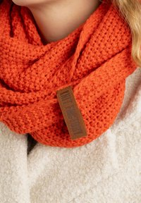 Een oranje gebreide sjaal met een textuurpatroon en een bruine leren label met de tekst "Knit Factory." Gedragen over een fluffy witte jas.
