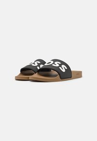 Des sandales noires à glissière présentent une large sangle avec un texte logo blanc. La semelle est beige, en caoutchouc, et a un design plat et décontracté.