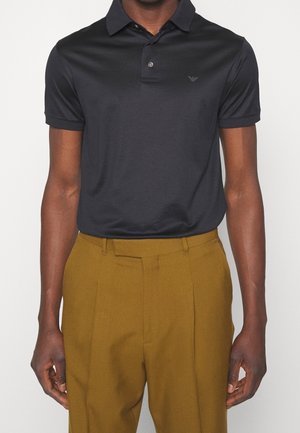 Homme portant un polo noir ajusté à manches courtes et un pantalon jaune moutarde taille haute, debout devant un fond uni.