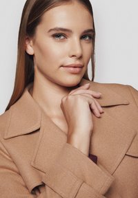 Jasno brązowy wełniany płaszcz z szerokimi klapami, zaprezentowany na osobie, która trzyma rękę w zamyśleniu blisko twarzy. Gładka tekstura, dopasowany krój.