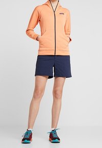 Sudadera naranja con cremallera y tejido texturizado, pantalones cortos azul marino y zapatillas deportivas multicolores. Presenta una capucha y bolsillos frontales.