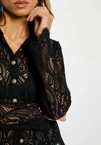 Blusa nera in pizzo con motivi intricati, chiusura frontale con bottoni e colletto morbido; presenta maniche lunghe con bordi sfrangiati e bottoni metallici.