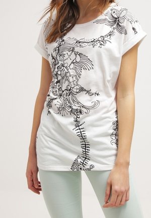 Frau trägt ein weißes Kurzarmshirt mit schwarzen abstrakten Blumen- und Linienmotiven und hellen Hosen und steht mit locker hängenden Armen.