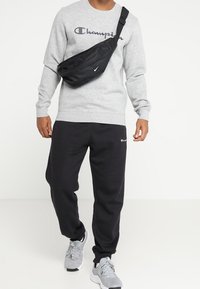 Felpa grigia con logo "Champion", borsa a tracolla nera, pantaloni sportivi neri e scarpe da ginnastica grigie. Outfit casual e sportivo con texture diverse.