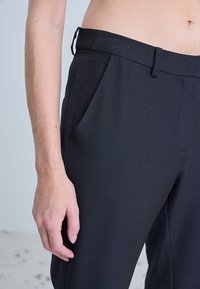 Gros plan sur une personne portant un pantalon noir taille haute avec des poches latérales et des passants pour ceinture, montrant la main gauche détendue sur le côté.