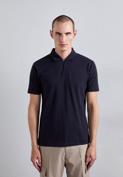 Polo shirt bleu marine à manches courtes, avec un col boutonné et un motif triangulaire à l'avant. Porté avec un pantalon cargo beige.