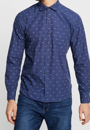 Marineblauw overhemd met knopen en lange mouwen, met een klein wit streepjespatroon, gedragen met donkerblauwe spijkerbroek, zichtbaar van borst tot heup.