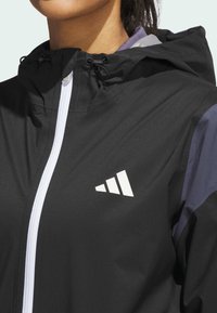 Black jacket with a white zipper and three-stripe logo on the chest; features a hood and textured fabric. Accented with purple details.