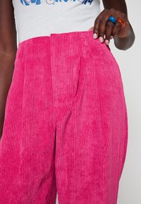 Pantalones de pana rosa con estrías verticales, que presentan un delantero con pliegues cuidados y una cinturilla ancha. Mano posicionada en la cintura.