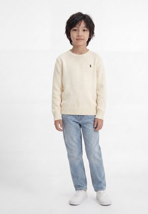 Garçon aux cheveux foncés portant un pull crème, un jean bleu clair et des baskets blanches, debout devant un fond blanc uni.