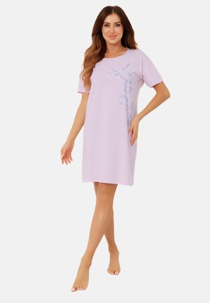 Kobieta o długich brązowych włosach, ubrana w jasnofioletową sukienkę do kolan z krótkimi rękawami i niebieskim wzorem liści, stojąca boso.