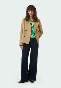 Beige double-breasted blazer with black buttons, worn over a green patterned top, paired with dark wide-leg denim, completed with black shoes.