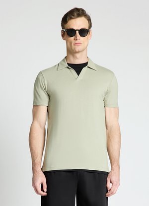 Uomo che indossa una polo verde chiaro a maniche corte, occhiali da sole neri e pantaloni neri, in piedi davanti a uno sfondo chiaro e uniforme.