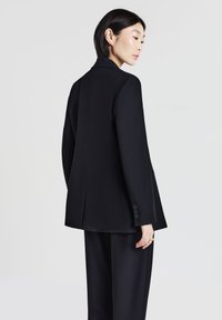 Blazer nero su misura con design strutturato, chiusura a un bottone e due bottoni su ciascun polsino. Tessuto liscio e testurizzato con una silhouette pulita.