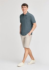 Camisa polo gris oscuro con collar y mangas cortas, combinada con pantalones cortos beige claros y zapatillas blancas. El modelo está de pie de manera casual con las manos en los bolsillos.