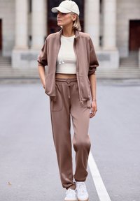 Beige zip-up jacket with a high collar, matching beige cropped top, and light brown tapered joggers. Worn with white sneakers and a cap.