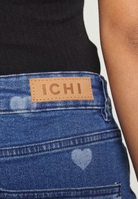 Blue denim jeans featuring a brown leather patch with "ICHI" embossed. Heart-shaped pattern on the fabric near the pocket. Smooth texture.