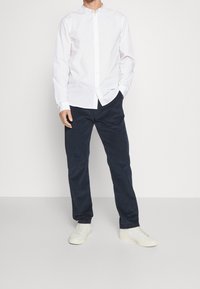 Camicia bianca con colletto a banda, abbinata a pantaloni blu navy e sneakers bianche. La camicia presenta maniche lunghe e una vestibilità slim.