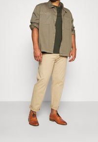 En kortärmad olivgrön skjorta över en mörkgrön t-shirt, kombinerad med beige byxor och bruna läderskor. Har två bröstfickor och uppvikta ärmar.