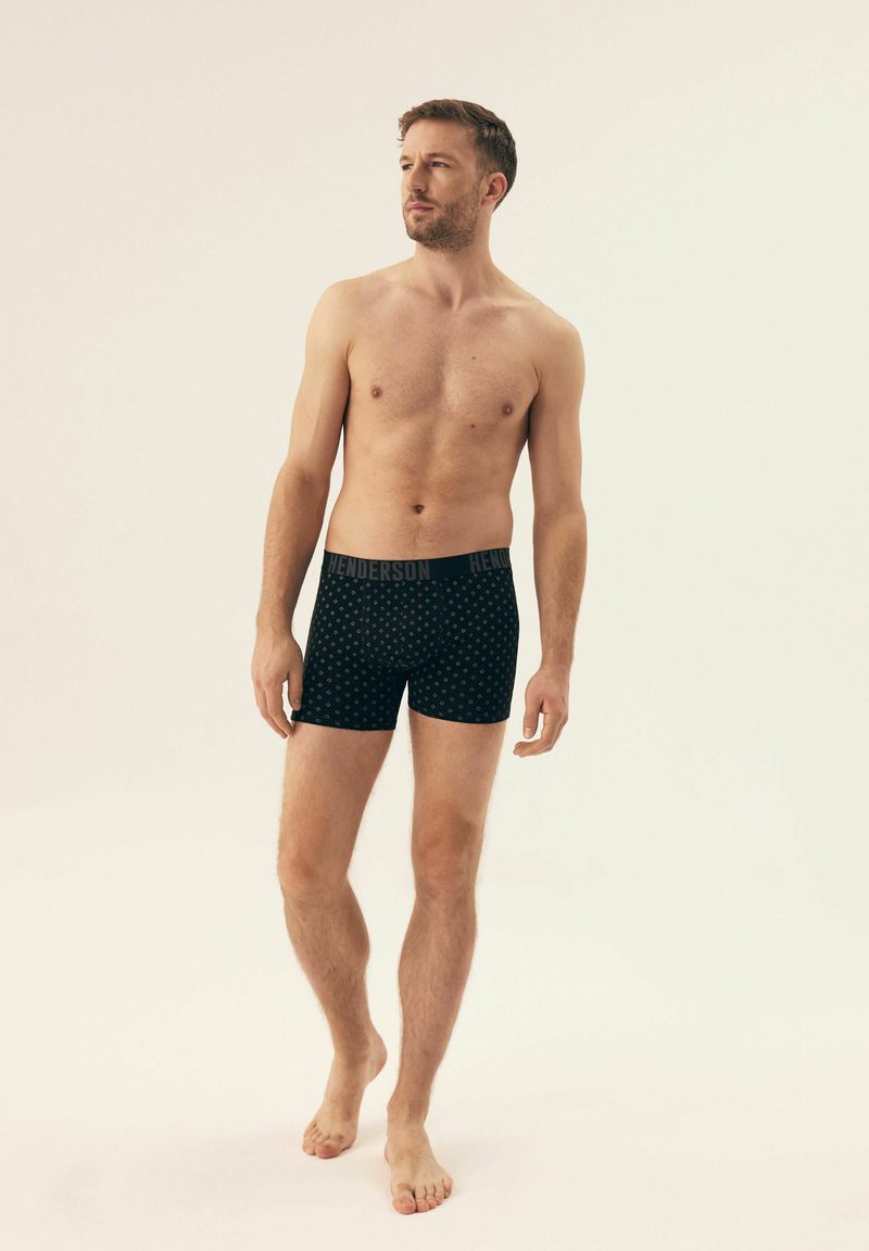 Schwarze Boxershorts mit einem dezenten Muster und einem gebrandeten Bund, getragen von einem Modell mit nacktem Oberkörper vor einem neutralen Hintergrund.
