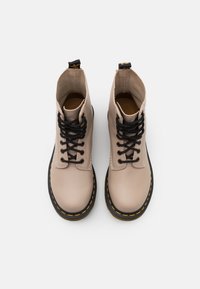 Botas de tobillo beige de cuero con cordones negros, un acento de costura amarilla y una suela de goma negra. Diseñadas con una punta redondeada.