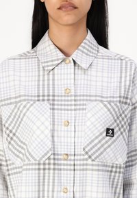 Camisa abotoada com padrão xadrez cinza e branco, tecido leve, dois bolsos frontais e um logótipo preto da Converse no bolso esquerdo.