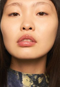 Nahaufnahme eines Gesichts mit glatter, heller Haut. Die Merkmale umfassen klare Augen, volle Lippen mit glänzendem Finish und glattes Haar. Der Hintergrund ist minimal.