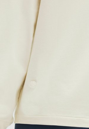 Krémszínű pulóver, puha anyagból készült, lekerekített aljjal és egy kicsi, hímzett logóval az oldalon.