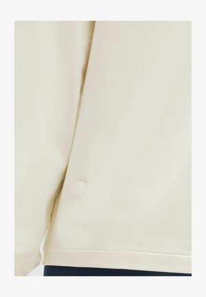 Krémszínű pulóver, puha anyagból készült, lekerekített aljjal és egy kicsi, hímzett logóval az oldalon.