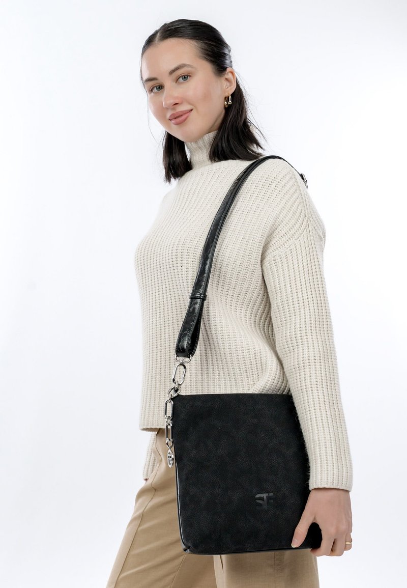 Femme portant un pull en maille beige et un pantalon marron clair, tenant un sac à bandoulière noir avec des éléments en argent, debout devant un fond blanc.