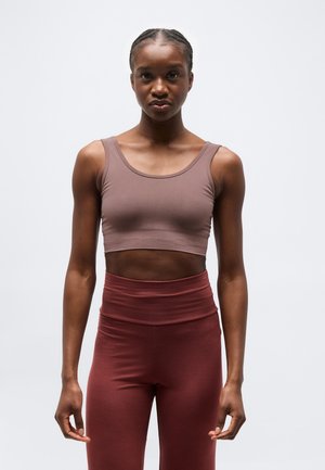 Fonott hajjal rendelkező nő barna crop topot és magas derekú bordó leggingset visel, fehér egyszínű háttér előtt áll.