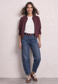 Femme aux cheveux mi-longs portant une veste grenat, une chemise blanche, un jean bleu et des baskets grenat, debout contre un mur uni.