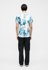 Άτομο με κοντά σκούρα μαλλιά, φορώντας λευκό T-shirt με μπλε κυματιστά σχέδια, μαύρο παντελόνι cargo και λευκά αθλητικά παπούτσια, που στέκεται με την πλάτη γυρισμένη.