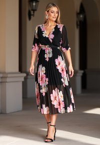 Czarna plisowana sukienka z różowo-białym kwiatowym wzorem, z krojem na kopertę, z rękawami 3/4 i paskiem. Noszona z czarnymi sandałami na obcasie.