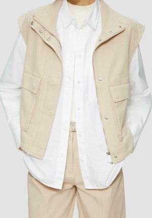 Gilet beige sans manches avec fermeture éclair et boutons-pression porté sur une chemise blanche à boutons et un pantalon beige. Mains dans les poches.
