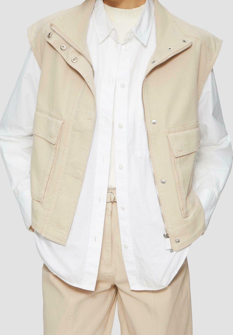 Gilet beige sans manches avec fermeture éclair et boutons-pression porté sur une chemise blanche à boutons et un pantalon beige. Mains dans les poches.