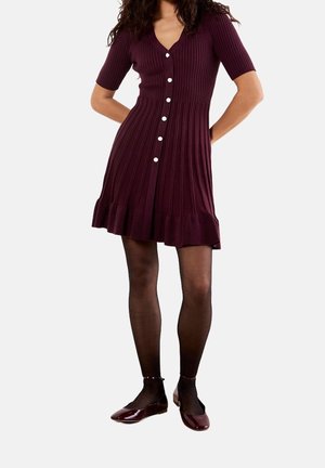 Mujer vistiendo un vestido burdeos de manga corta y canalé con botones blancos, medias negras transparentes y zapatos planos burdeos brillantes, de pie con las manos atrás.