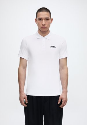 Vīrietis stāv ar seju uz priekšu, valkājot baltu īsroku polo krekliņu ar uzrakstu "Karl Lagerfeld" uz krūtīm un melnas bikses.