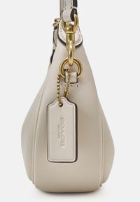 Vue latérale d’un sac à main Coach en cuir beige avec des ferrures dorées et une étiquette en cuir assortie suspendue à une chaîne à billes dorée.
