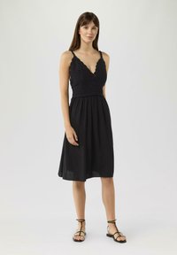 Schwarzes Kleid mit einem enganliegendem Strick-Oberteil und einem ausgestellten Rock, mit dünnen verstellbaren Trägern und einem V-Ausschnitt. Ein Paar schwarze flache Sandalen.