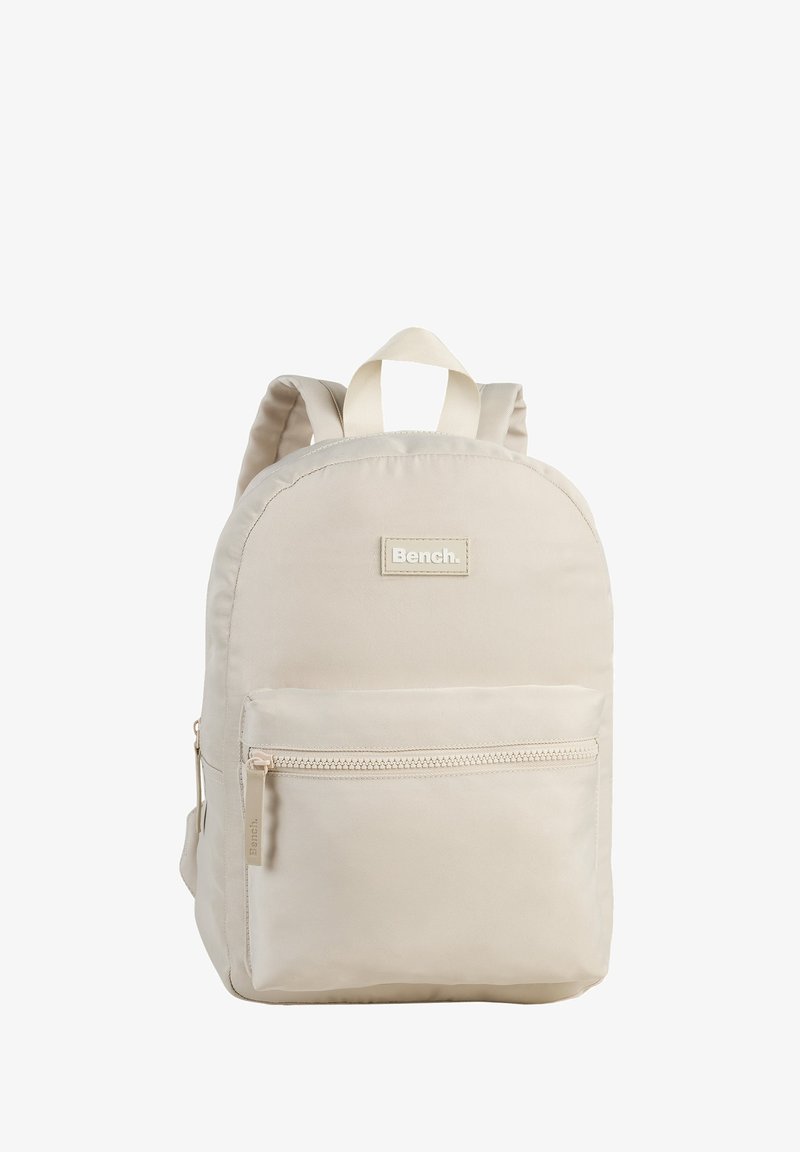 Beige Stoffrucksack mit runder Form, vorderer Reißverschlusstasche, Logo-Aufnäher und zwei Schulterriemen. Glatte Textur und minimalistisches Design.
