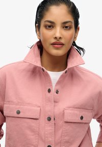 Veste en toile rose avec un col haut, deux poches avant et des boutons noirs. Texture lisse et coupe cintrée.