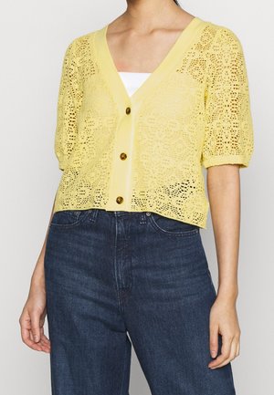Kvinde iført en gul blonder kortærmet cardigan med knapper over en hvid top, kombineret med højtaljede mørkeblå jeans.