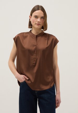 Mujer con cabello corto castaño, vistiendo una blusa marrón sin mangas con botones y jeans azul oscuro, de pie con una mano en el bolsillo.