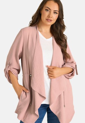Femme portant un cardigan rose pâle ouvert à l'avant avec les manches retroussées, sur une chemise blanche et un jean bleu, posant avec une main dans la poche.