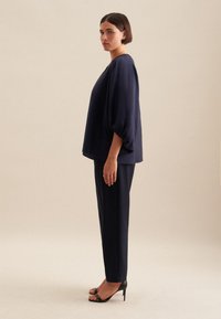 Blusa blu navy con maniche ampie, abbinata a pantaloni blu navy aderenti e tacchi neri con cinturini. Tessuto liscio, design semplice, nessun motivo visibile.