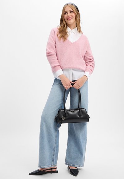 Femme portant un pull rose sur une chemise blanche avec un jean à jambes larges et des chaussures noires pointues, tenant un sac à main noir avec deux poignées.