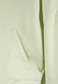 Ljust grön jacka med en slät, glänsande yta, med en sidoficka och en modern, minimalistisk design. Inga synliga beslag eller mönster.