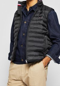 Czarna kamizelka puchowa z pikowanym wzorem, wysokim kołnierzem i przednim zamkiem błyskawicznym. Nosi się ją na koszulę dżinsową, z widocznymi elementami metalowymi i warstwową strukturą.